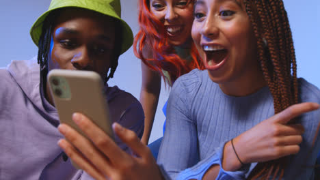 Close-Up-Studio-Shot-Of-Young-Gen-Z-Friends-Sitting-On-Sofa-Sharing-Social-Media-Post-On-Mobile-Phones